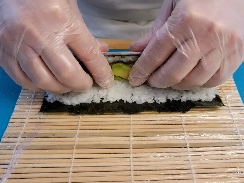Mains gantées roulant un maki à l'avocat sur natte